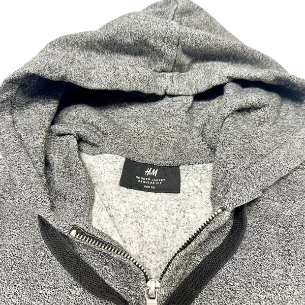 Gros plan sur la capuche grise d’un sweat zippé H&M, tissu chiné et étiquette visible à l’intérieur.