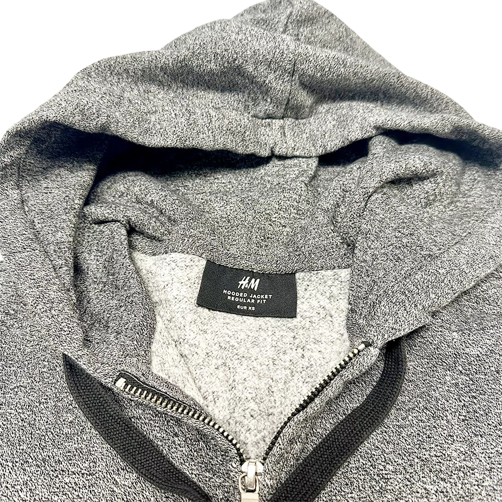 Gros plan sur la capuche grise d’un sweat zippé H&M, tissu chiné et étiquette visible à l’intérieur.