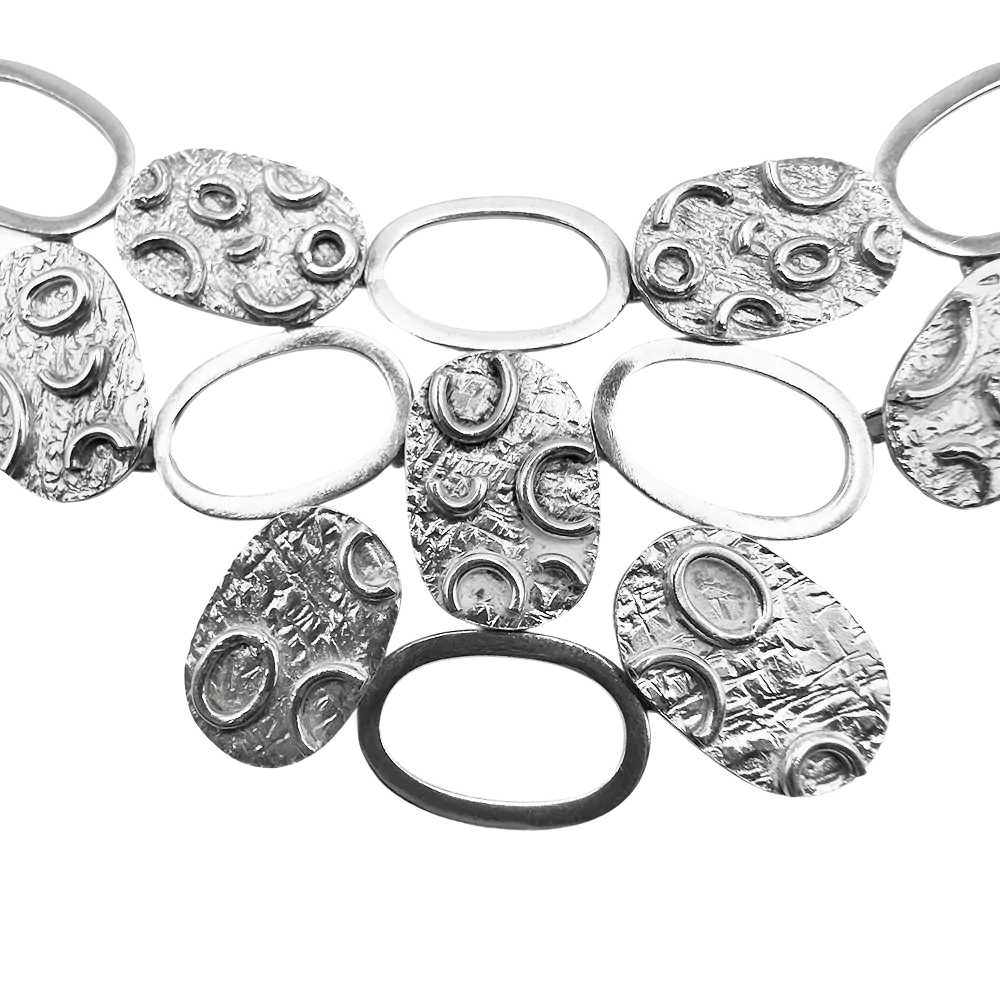 Srebrna ogrlica z ovalnimi diski in teksturiranim vzorcem, razstavljena na belem ozadju.